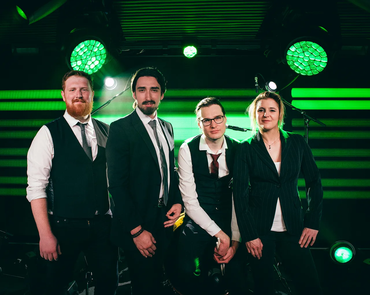 Four people dressed in formal attire standing and sitting on a stage with green lighting effects in the background.