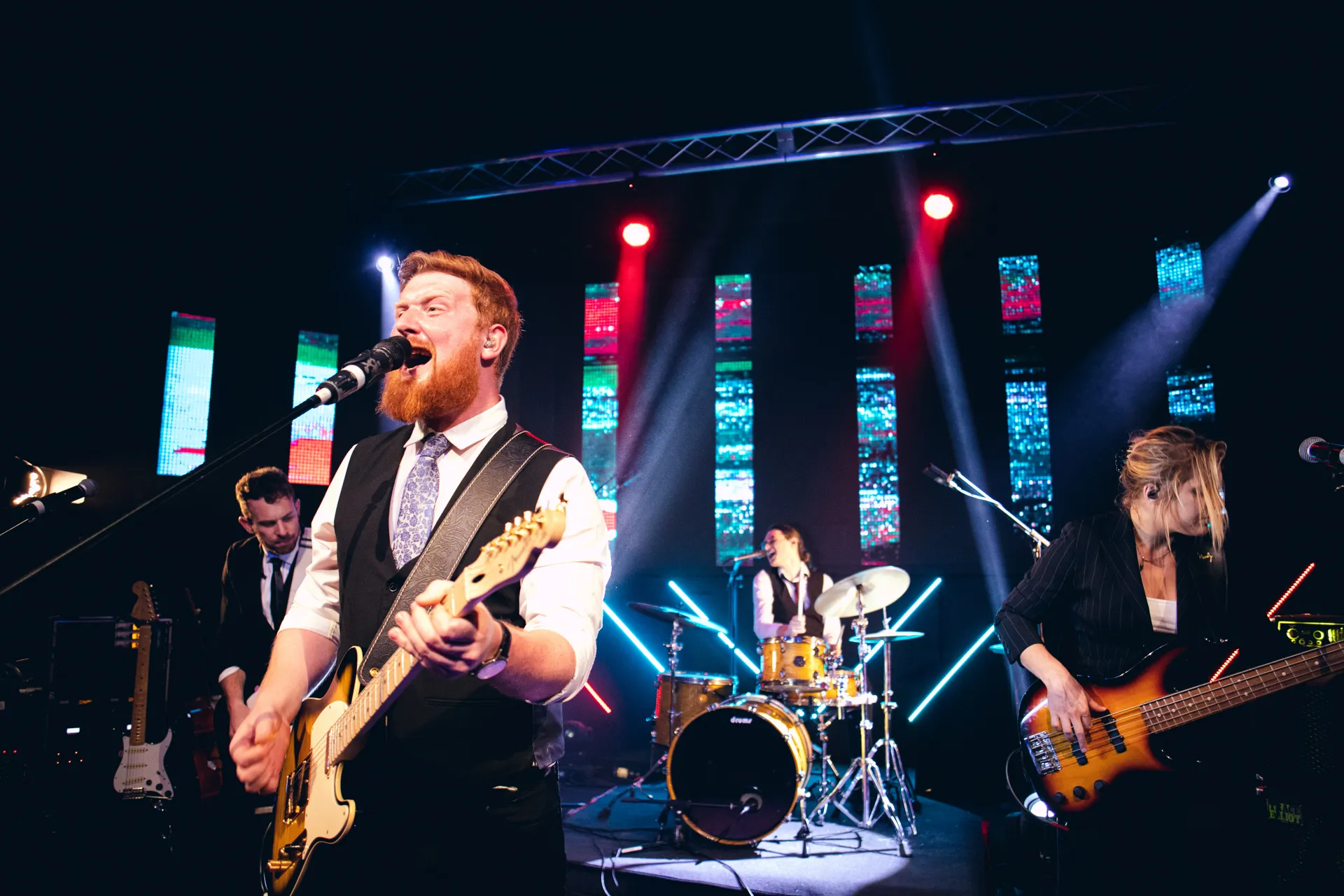 Four-piece band performing on stage with colorful LED lights in the background, featuring a male guitarist singing and a female bassist.