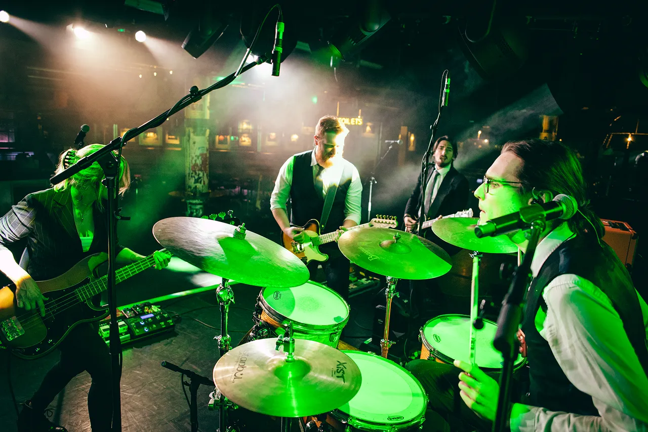Four-piece band performing on stage with drums, electric guitars, and bass under green stage lighting.