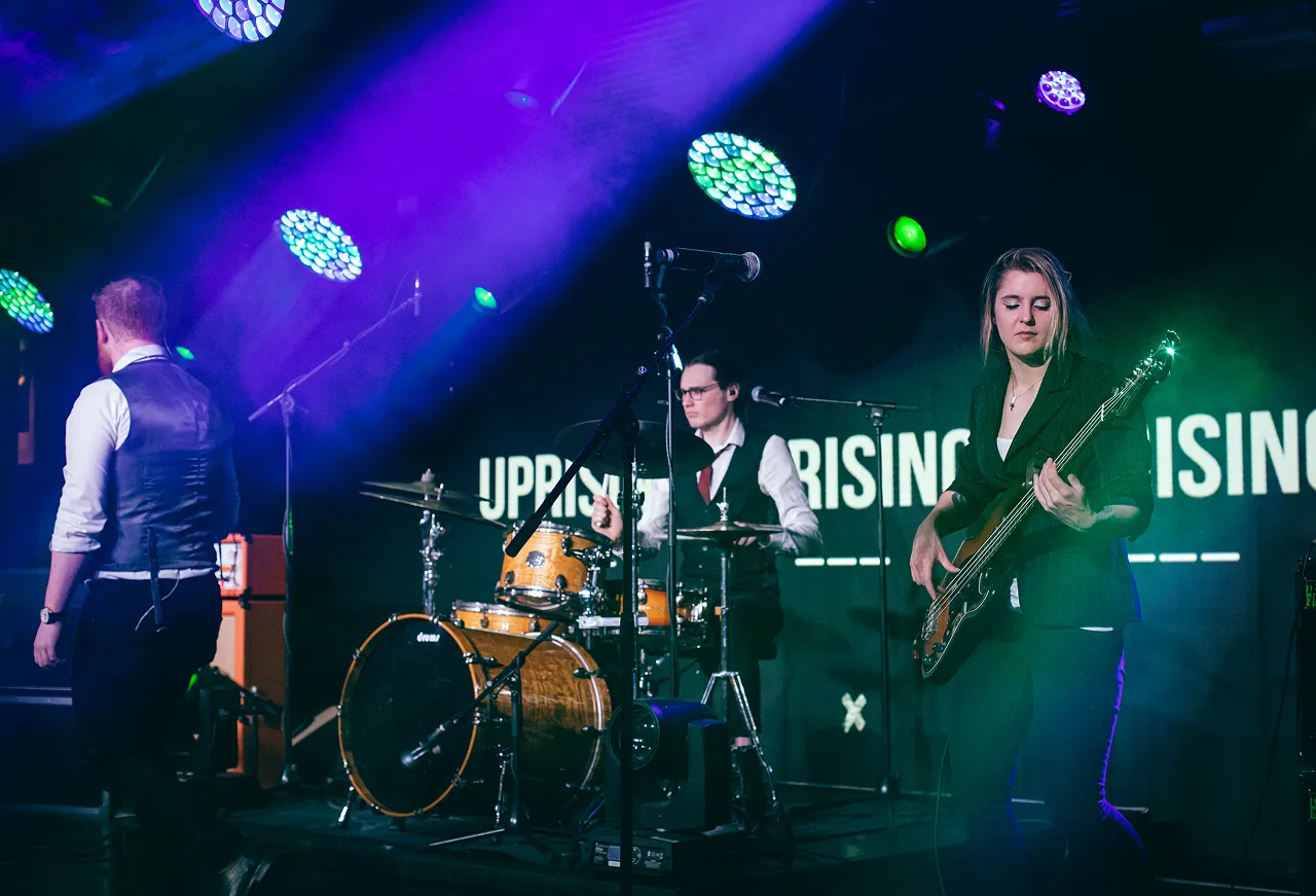 Band performing on stage with a drummer, bassist, and lead singer under purple and green stage lights.