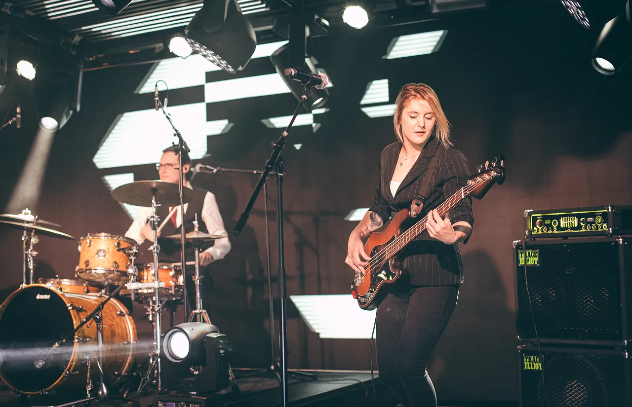 Female musician playing electric bass guitar on stage with drummer in the background under stage lights.