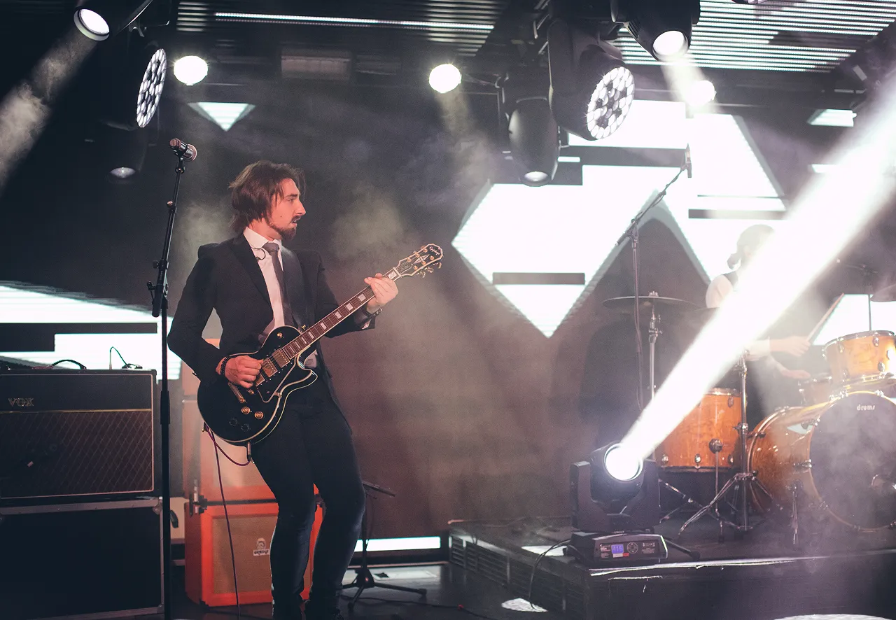 Man in a suit playing electric guitar on stage with drums and bright spotlights.
