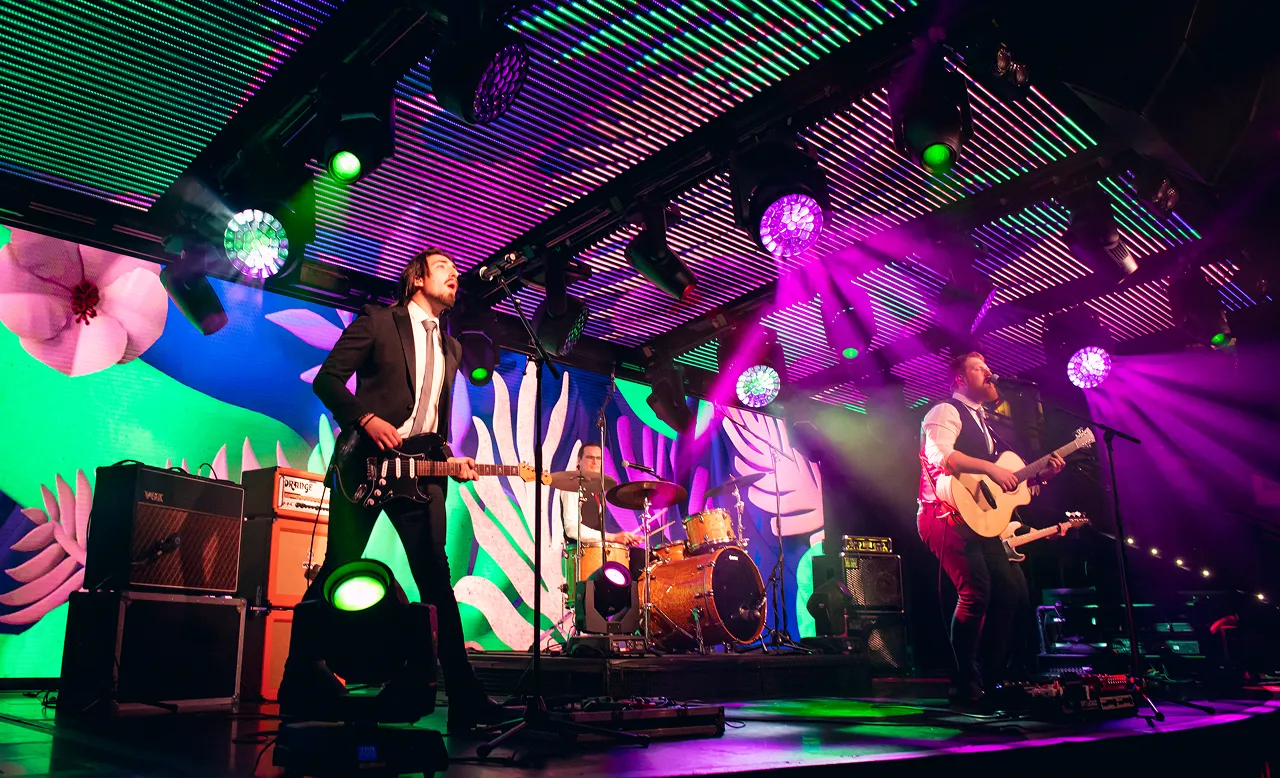 Band performing on stage with electric guitar, acoustic guitar, and drums under vibrant purple and green stage lights and colorful floral backdrop.